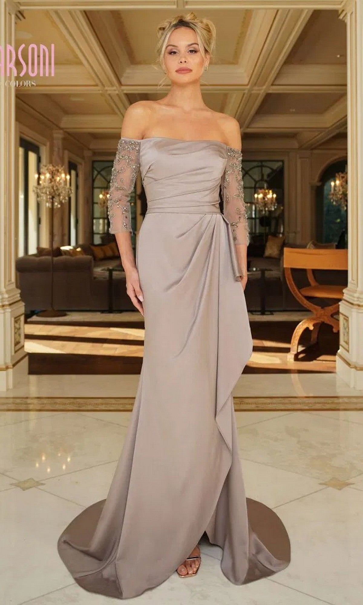 A woman stands indoors in a well-lit, elegant room with chandeliers, wearing the Marsoni MV1254 long formal dress in taupe, featuring off-the-shoulder sheer beaded sleeves and a draped skirt.